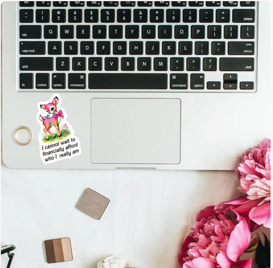 A die-cut sticker featuring a cartoon pink fox wearing a dress and holding a bag, with the text 'I cannot wait to financially afford who i really am' below it. The sticker is placed on a white laptop keyboard, styled with a gold ring, makeup sponges, and pink flowers on a light surface.