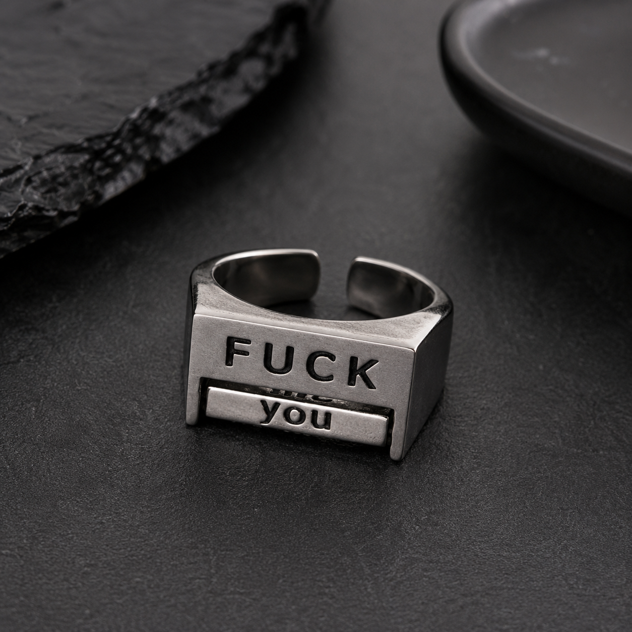 A silver-toned metal ring with a rectangular face engraved with the words 'FUCK you' in black lettering, resting on a dark textured surface.