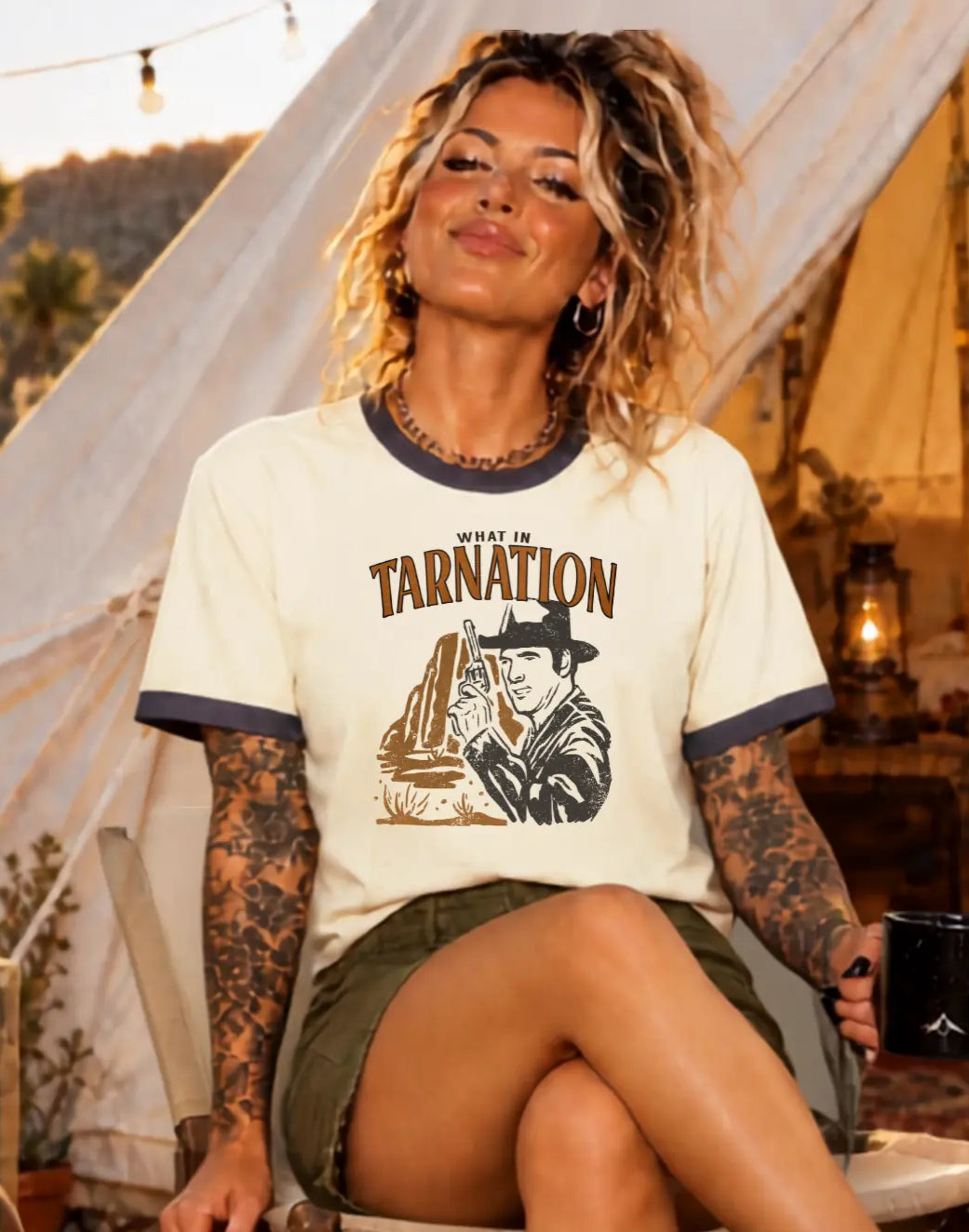 A woman sits on a wooden chair in a rustic campsite setting with a canvas tent, lanterns, and a patterned rug. She is wearing a cream-colored graphic t-shirt with dark blue trim and the text 'WILD WEST OF TARNATION' with a cowboy illustration, olive green shorts, and leopard-print sneakers.