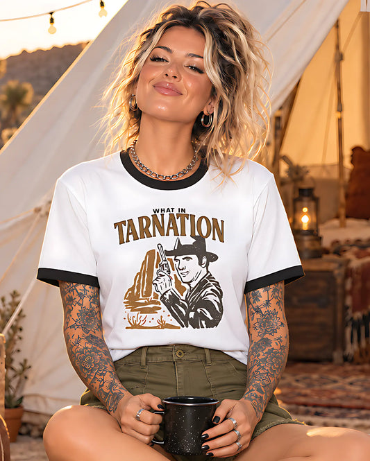 A woman wearing a white ringer t-shirt with black trim and a graphic print that reads 'WHAT IN TARNATION' with an illustration of a cowboy and a desert landscape. She is holding a black mug in a warm, rustic outdoor setting with a canvas tent in the background.