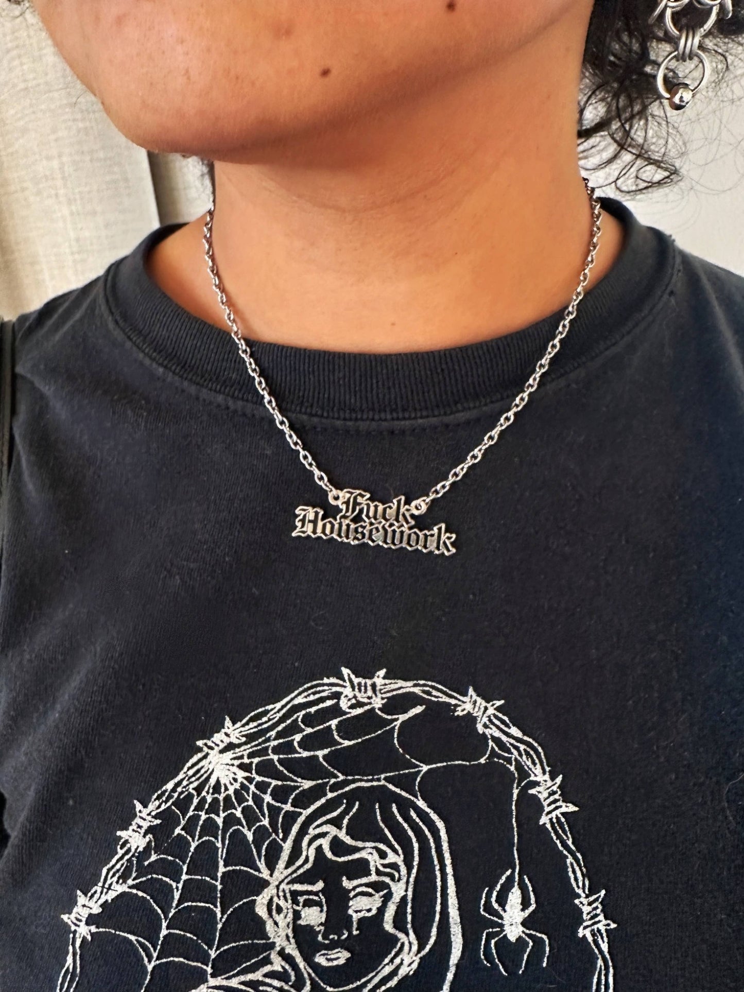 A close-up of a person wearing a silver gothic-style chain necklace with the words 'Fuck Housework' in a high-contrast font, paired with a black graphic t-shirt.