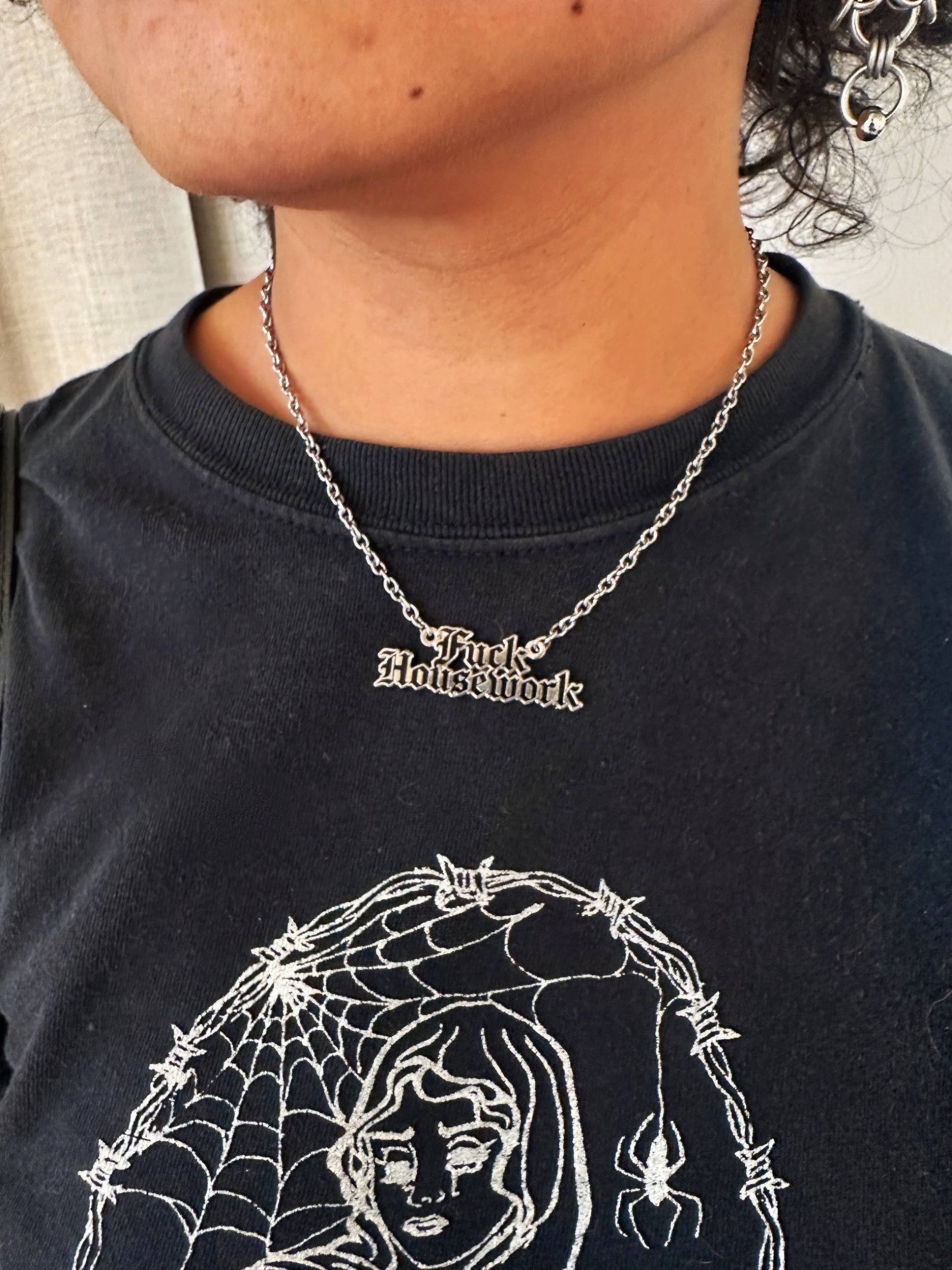A close-up of a person wearing a silver gothic-style chain necklace with the words 'Fuck Housework' in a high-contrast font, paired with a black graphic t-shirt.