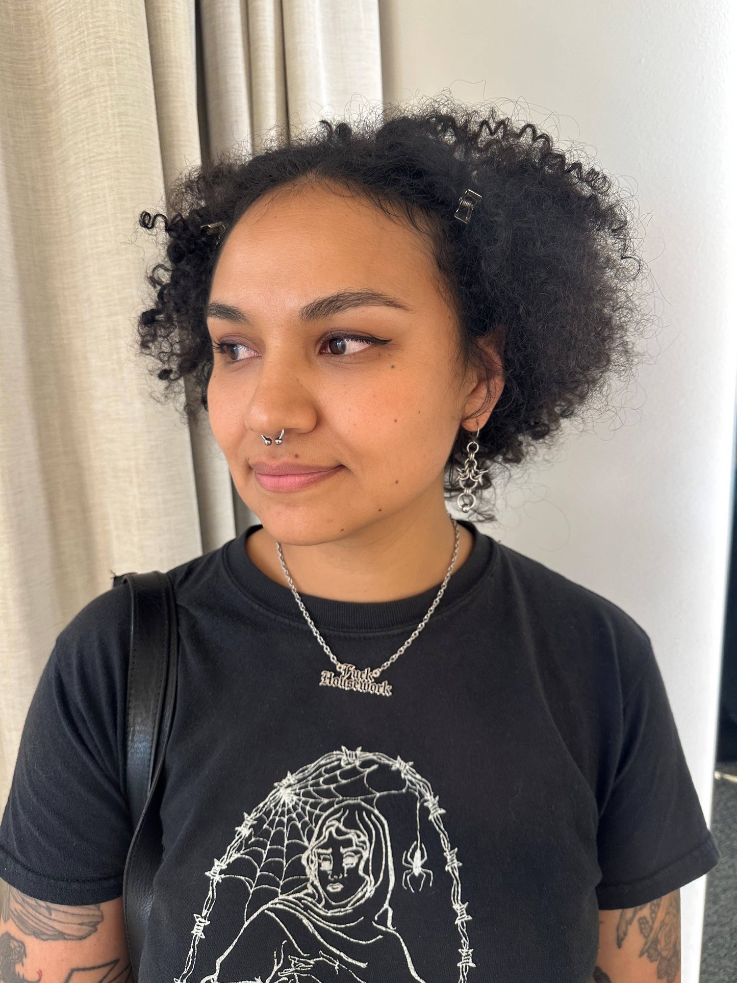 A woman with curly black hair wearing a black graphic t-shirt with a white spider-themed illustration, a silver necklace, and a nose ring, standing indoors in front of a curtain.