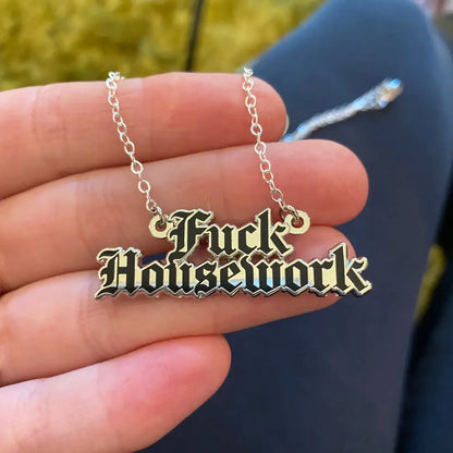 A close-up of a hand holding a silver chain necklace with a gothic-style metal pendant that reads 'Fuck Housework' in raised letters with a black outline and gold fill.