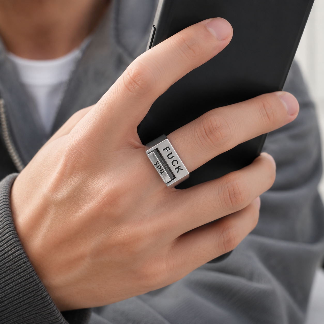 A close-up of a person's hand holding a black smartphone, wearing a silver rectangular ring engraved with the words 'FUCK YOU'.
