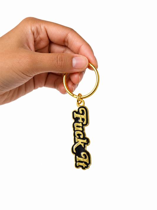 A hand holding a gold-tone keychain with a black and gold 'Fuck It' script charm against a solid white background.