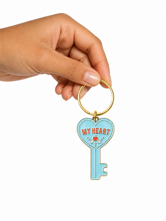 A hand holding a light blue key-shaped charm with a gold metal key ring. The charm features the words 'MY HEART' in orange with a small red heart and white decorative details.