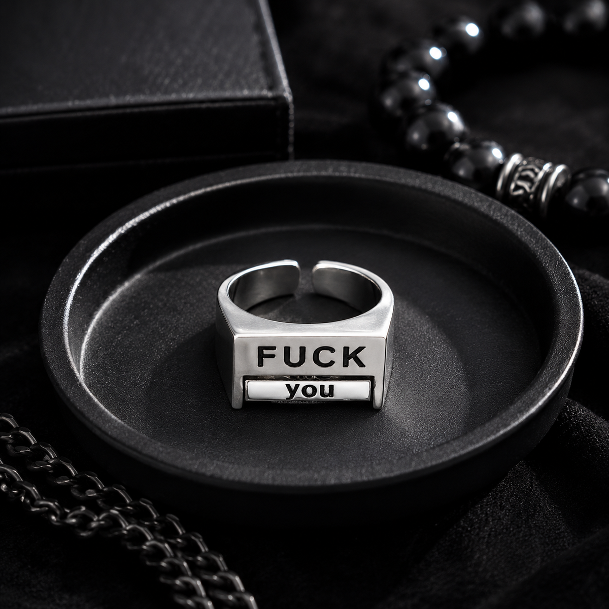 A silver-toned metal ring displayed in a black oval tray, featuring the engraved text 'FUCK YOU' on its front. The scene is styled with black jewelry accessories and a black box in the background.