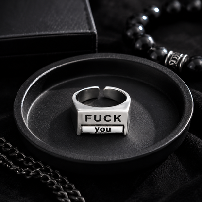 A silver-toned metal ring displayed in a black oval tray, featuring the engraved text 'FUCK YOU' on its front. The scene is styled with black jewelry accessories and a black box in the background.