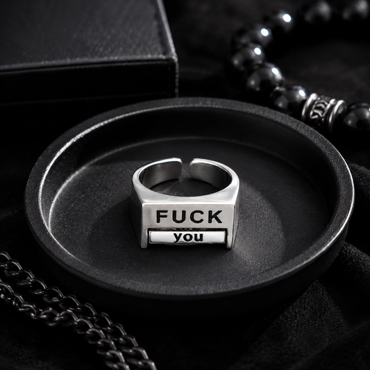 A silver-toned metal ring displayed in a black oval tray, featuring the engraved text 'FUCK YOU' on its front. The scene is styled with black jewelry accessories and a black box in the background.