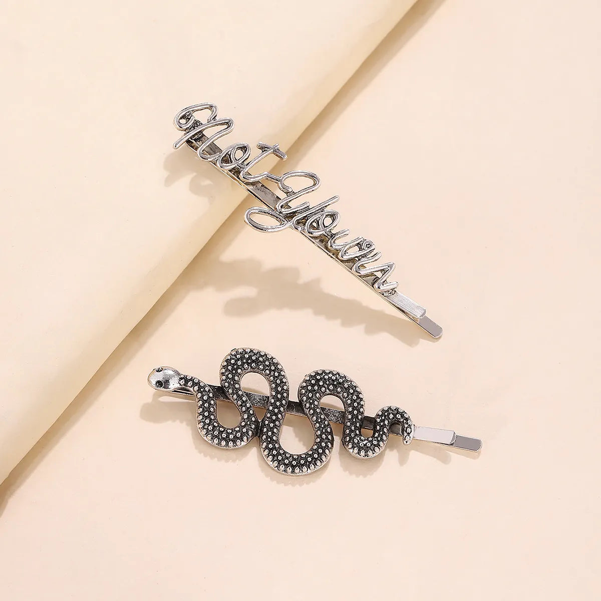 Two silver hair claw clips styled on a beige background. One clip features the word 'motivation' in cursive script, and the other is a coiled snake design with textured black and silver scales.