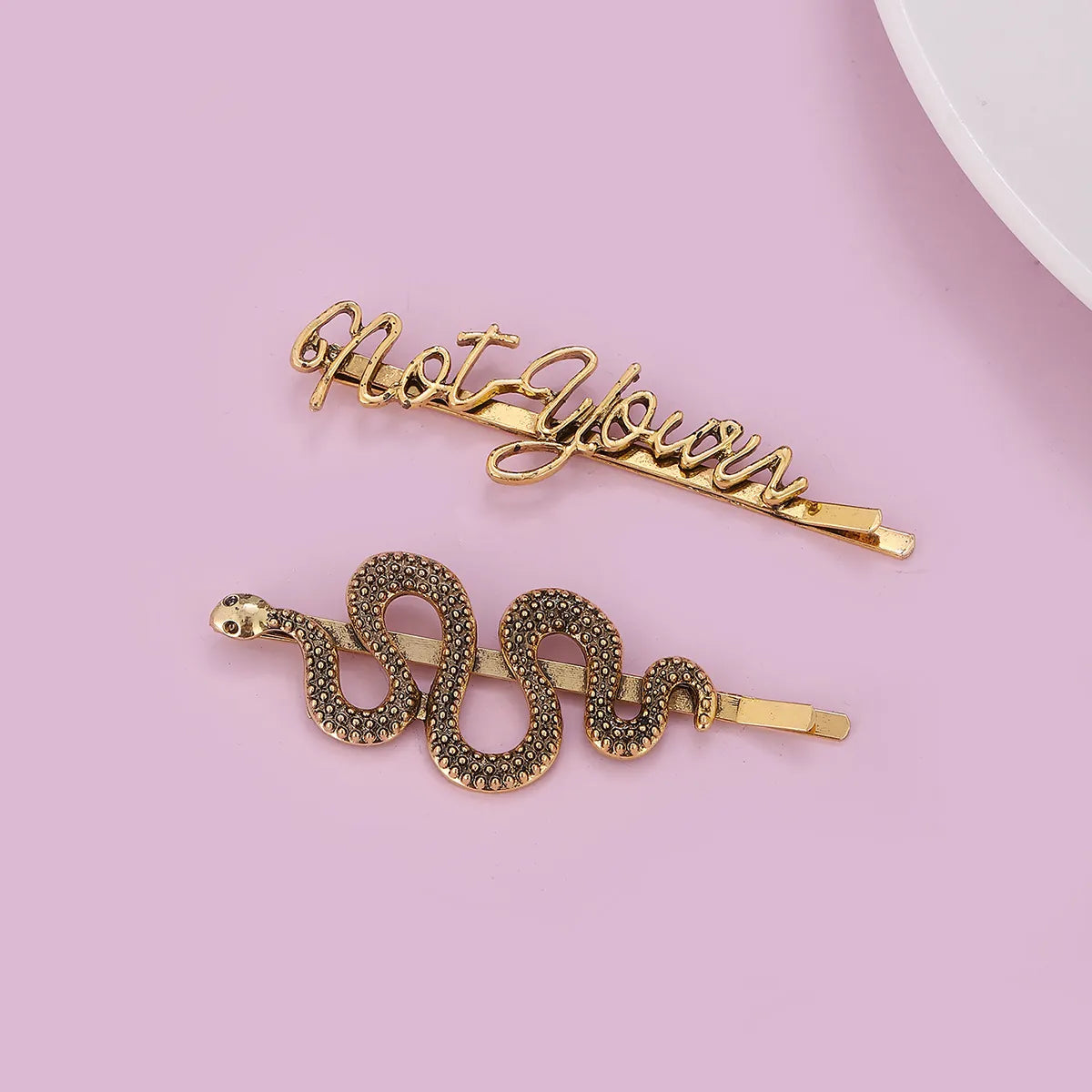 Two gold-toned hair barrettes on a light pink background: one with cursive text reading 'Not Yours' and one shaped like a coiled snake covered in small dark rhinestones.