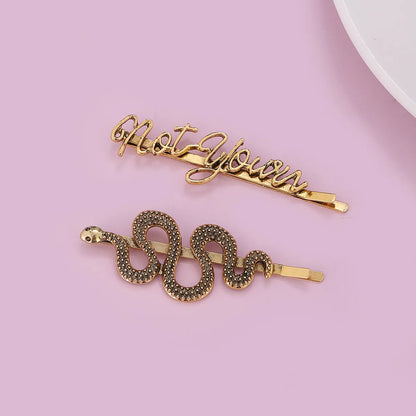 Two gold-toned hair barrettes on a light pink background: one with cursive text reading 'Not Yours' and one shaped like a coiled snake covered in small dark rhinestones.
