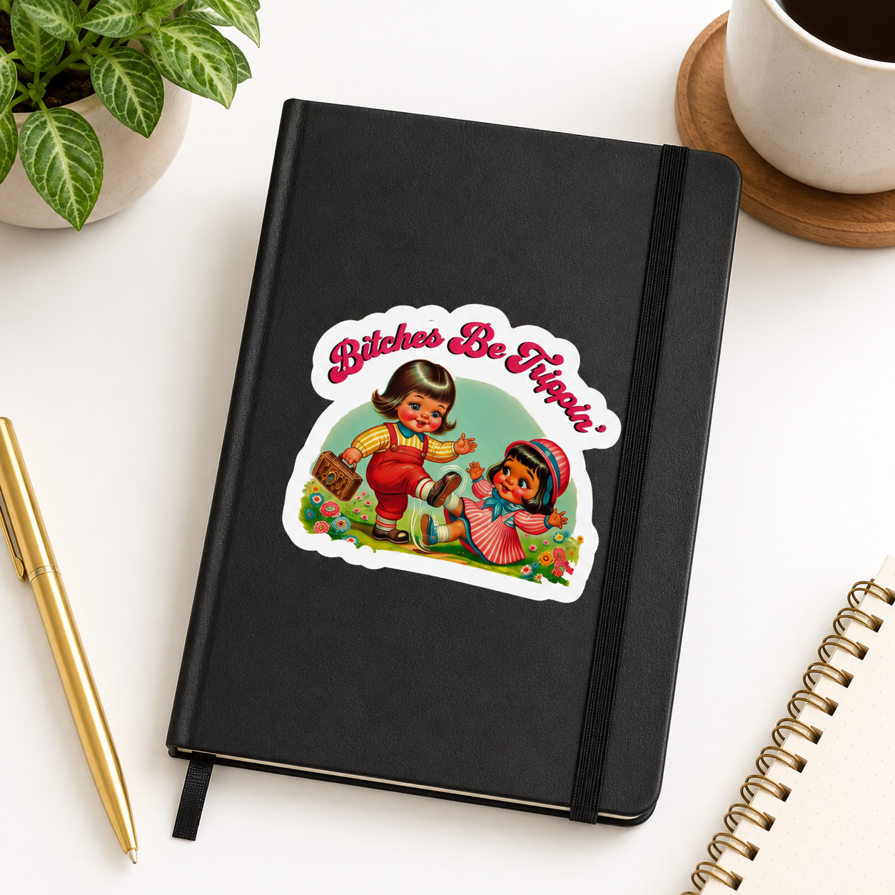 A black notebook with an elastic closure and a die-cut sticker on the cover that reads 'Bitches Be Hip!' with a retro illustration of two children. The notebook is photographed on a white desk with a plant, coffee cup, pen, and spiral notebook around it.