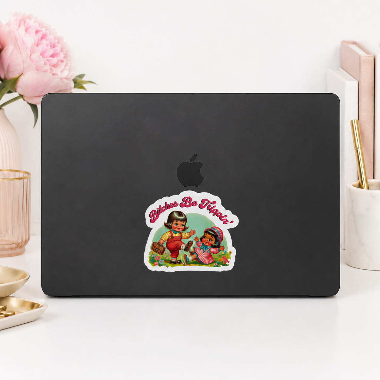 A black laptop with a colorful retro-style sticker on the back, styled on a white desk with pink flowers and stationery in the background.