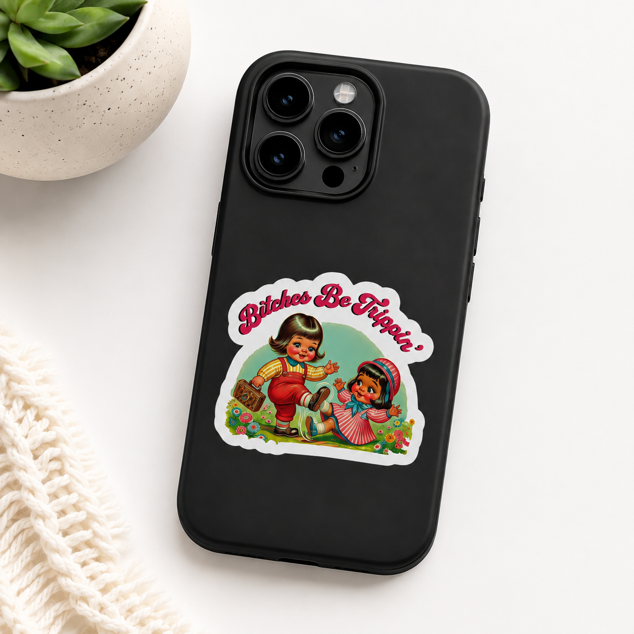 A black phone case featuring a retro-style die-cut sticker with the text 'Bitches Be Hippie' and an illustration of two women in colorful clothing, shown on a white background with a small potted succulent and cream knit fabric as props.