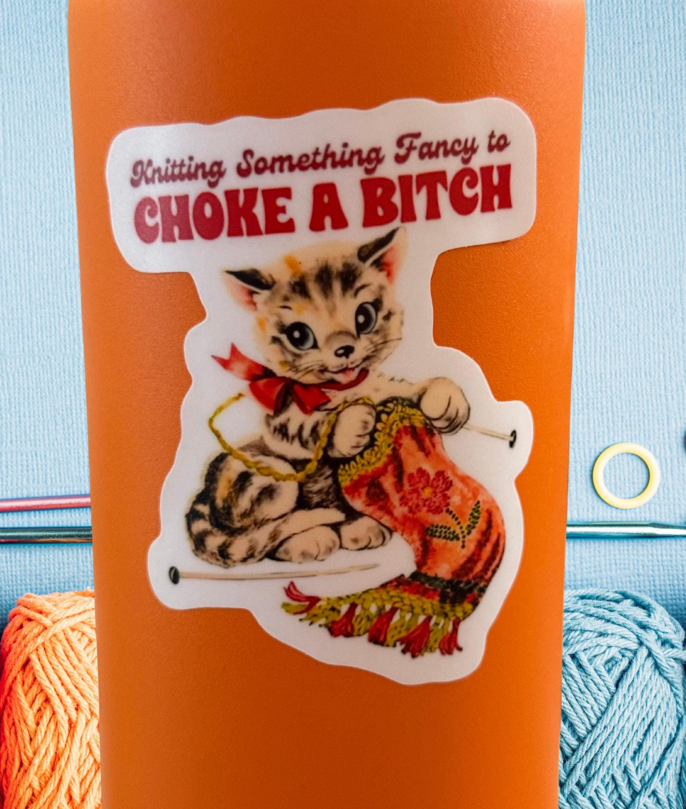 An orange insulated tumbler with a die-cut sticker featuring a tabby kitten knitting a red and orange patterned scarf, with the text 'Knitting Something Fancy to CHOKE A BITCH' in red and black lettering. The tumbler is photographed against a light blue background with knitting yarn visible at the bottom corners.