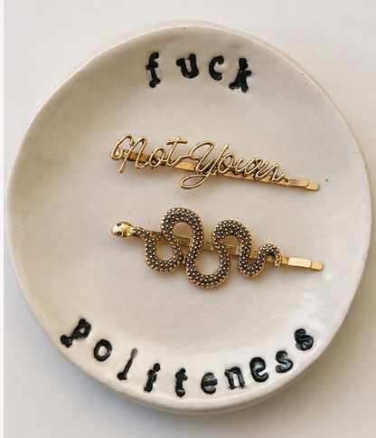 Two decorative white ceramic dishes with the words 'fuck politeness' in black lettering, each holding a coiled snake-shaped hair pin in different metal finishes: one gold-toned and one silver-toned. The dishes are shown side by side against a plain light background.