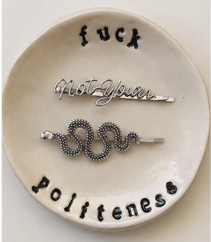 Decorative white ceramic dish with the words 'fuck politeness' in black lettering, each holding a coiled snake-shaped hairpin in different metal finishes: one gold-toned and one silver-toned. The dishes are shown side by side against a plain light background.
