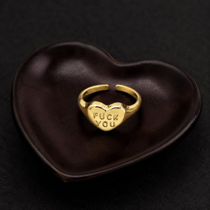 A gold heart-shaped ring engraved with the words 'FUCK YOU' sits in a dark brown heart-shaped dish on a black background.