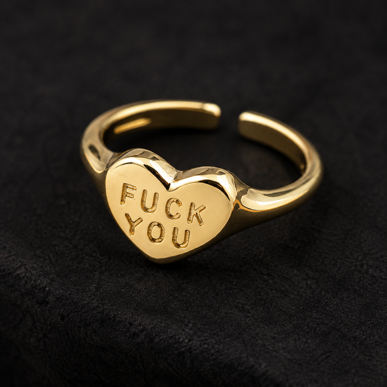 A gold-plated open band ring featuring a heart-shaped face with the words 'FUCK YOU' engraved on it, set against a dark textured background.