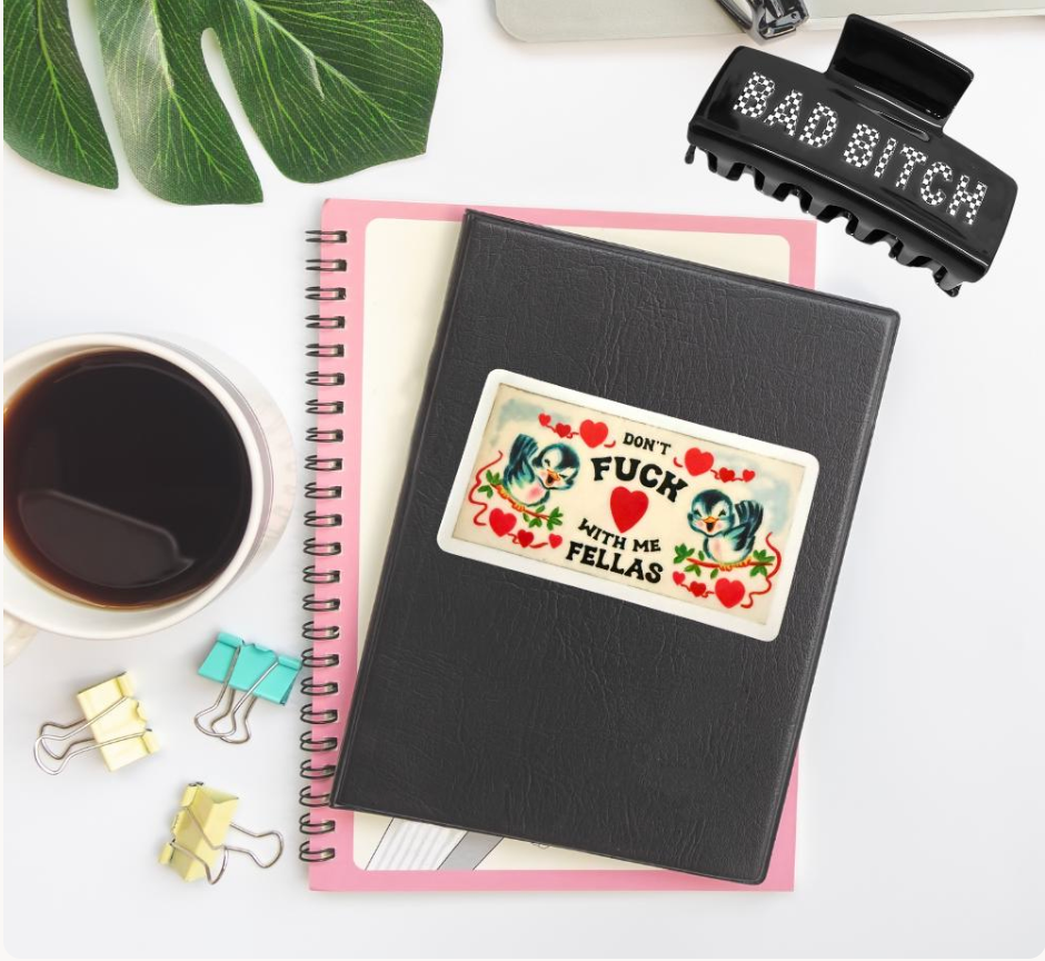 A black spiral notebook with a decorative sticker on the cover, styled in a flat-lay arrangement with a pink page insert, a cup of black coffee, binder clips, a green leaf, and a black hair claw clip on a white background.