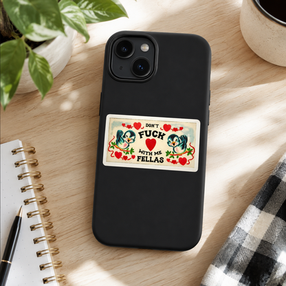 A black phone case with a rectangular graphic sticker on the back, displayed on a light wood desk with a notebook, plant, and coffee cup nearby.