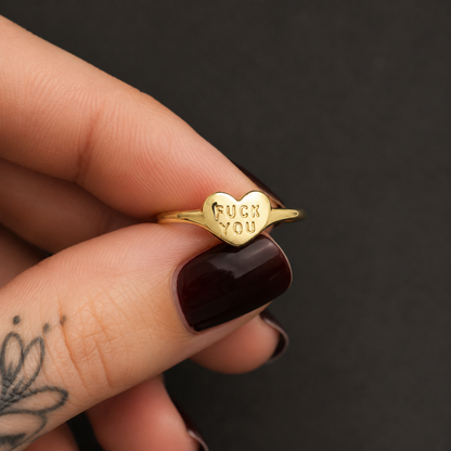 A gold ring with a heart-shaped center engraved with the words 'FUCK YOU', held between fingers with dark burgundy nail polish against a dark gray background.