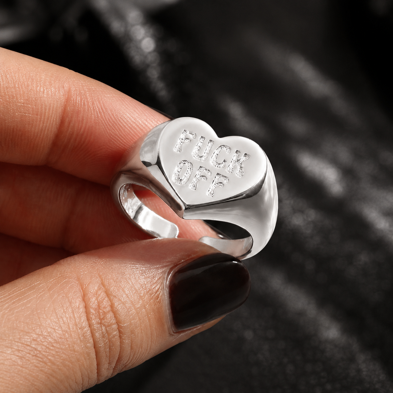 A close-up of a hand holding a silver signet ring with a heart-shaped face engraved with the words 'FUCK OFF'. The hand has dark brown nail polish, and the background is a dark, textured black fabric.