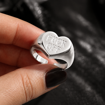 A close-up of a hand holding a silver signet ring with a heart-shaped face engraved with the words 'FUCK OFF'. The hand has dark brown nail polish, and the background is a dark, textured black fabric.