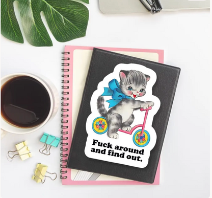 A black spiral notebook with a die-cut sticker of a gray kitten riding a pink bicycle, placed on top of a pink notebook. The scene is styled as a flat lay with a cup of coffee, binder clips, a green leaf, and a pen on a white desk.