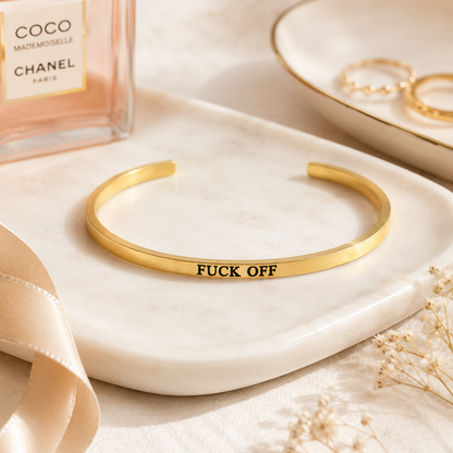 A gold cuff bracelet engraved with the text 'FUCK OFF' in black lettering, displayed on a white marble tray next to a Chanel perfume bottle and gold rings.