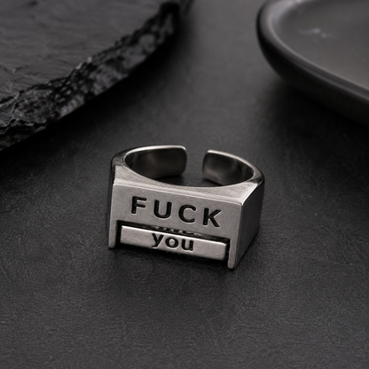 A silver-toned metal ring with a rectangular face engraved with the words 'FUCK you' in black lettering, resting on a dark textured surface.