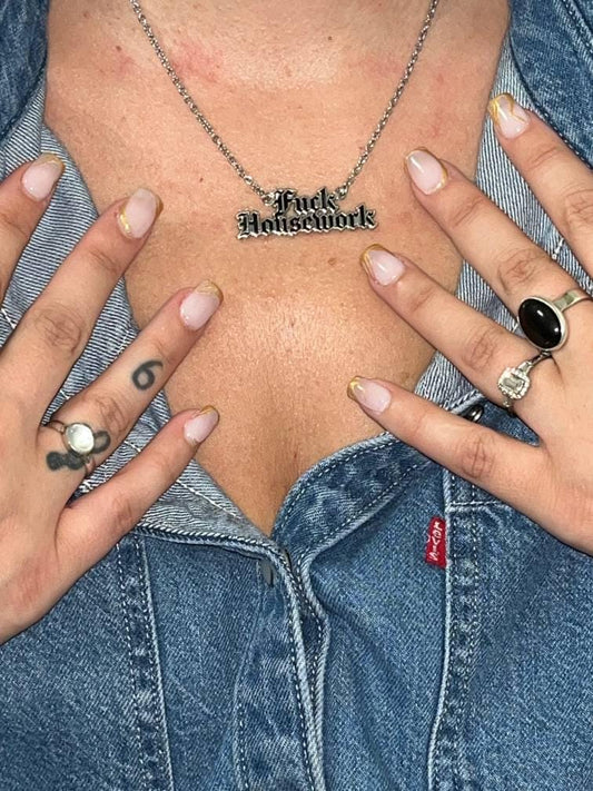 A close-up lifestyle shot of a person wearing a silver chain necklace with a 'Fuck Housework' pendant, styled with a denim shirt and multiple rings.