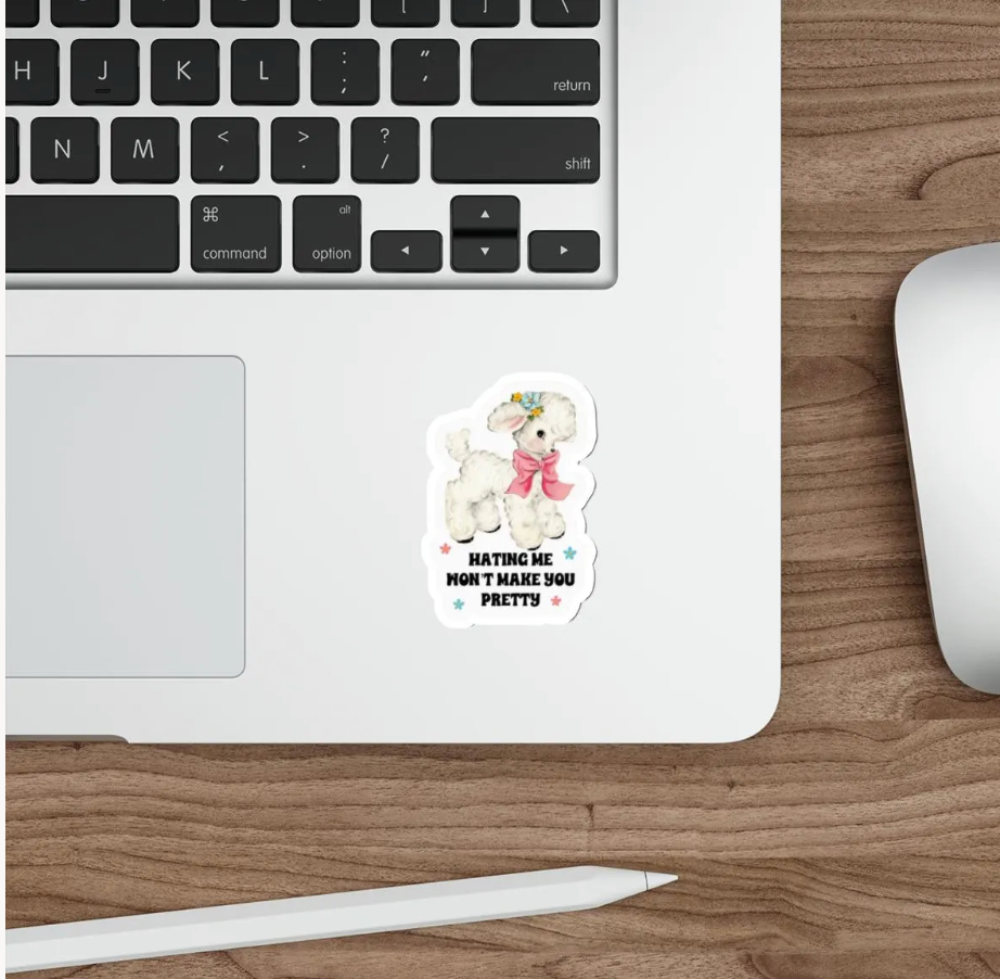 A die-cut sticker featuring a cute white poodle wearing a pink bow and gold crown, with the text 'HATING ME WON'T MAKE YOU PRETTY' in black and white, shown applied to the bottom corner of a silver laptop on a wooden desk next to a mouse and stylus.