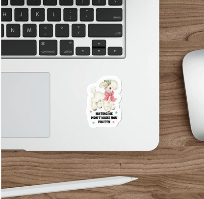 A die-cut sticker featuring a cute white poodle wearing a pink bow and gold crown, with the text 'HATING ME WON'T MAKE YOU PRETTY' in black and white, shown applied to the bottom corner of a silver laptop on a wooden desk next to a mouse and stylus.