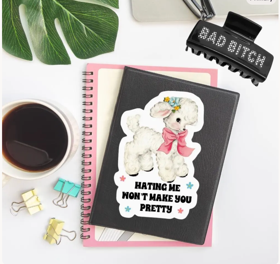 A black spiral notebook with a die-cut sticker of a fluffy white poodle wearing a pink bow and the text 'HATING ME WON'T MAKE YOU PRETTY' is styled on a white desk with a pink notebook underneath, a coffee cup, binder clips, a hair clip, and a green leaf.