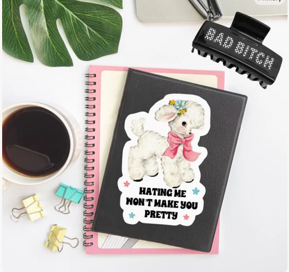 A black spiral notebook with a die-cut sticker of a fluffy white poodle wearing a pink bow and the text 'HATING ME WON'T MAKE YOU PRETTY' is styled on a white desk with a pink notebook underneath, a coffee cup, binder clips, a hair clip, and a green leaf.