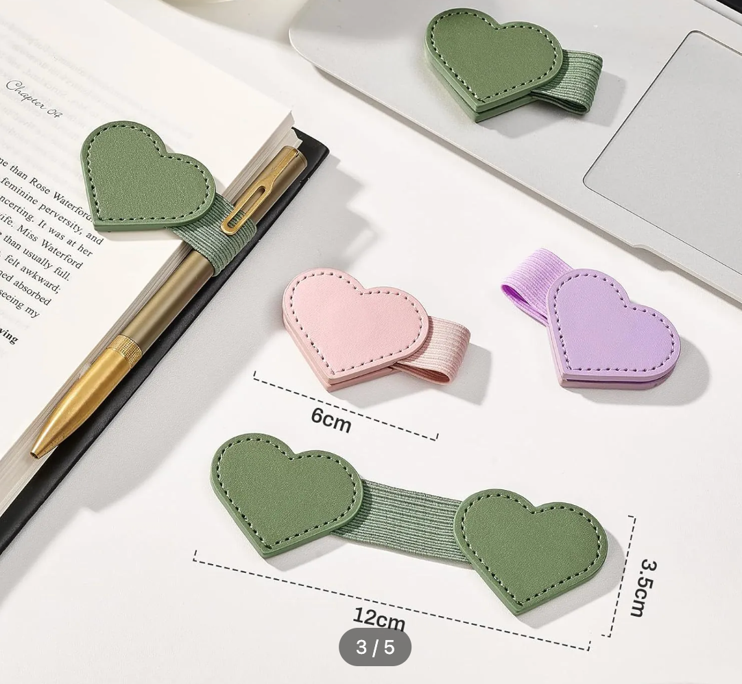 Heart-shaped elastic desk accessories shown on a white surface with overlaid dimension labels. The set includes a green pen holder, a pink notebook clip, a purple hair tie, and a larger green desk mat with heart ends, with one matching green heart clip also shown near the top right.