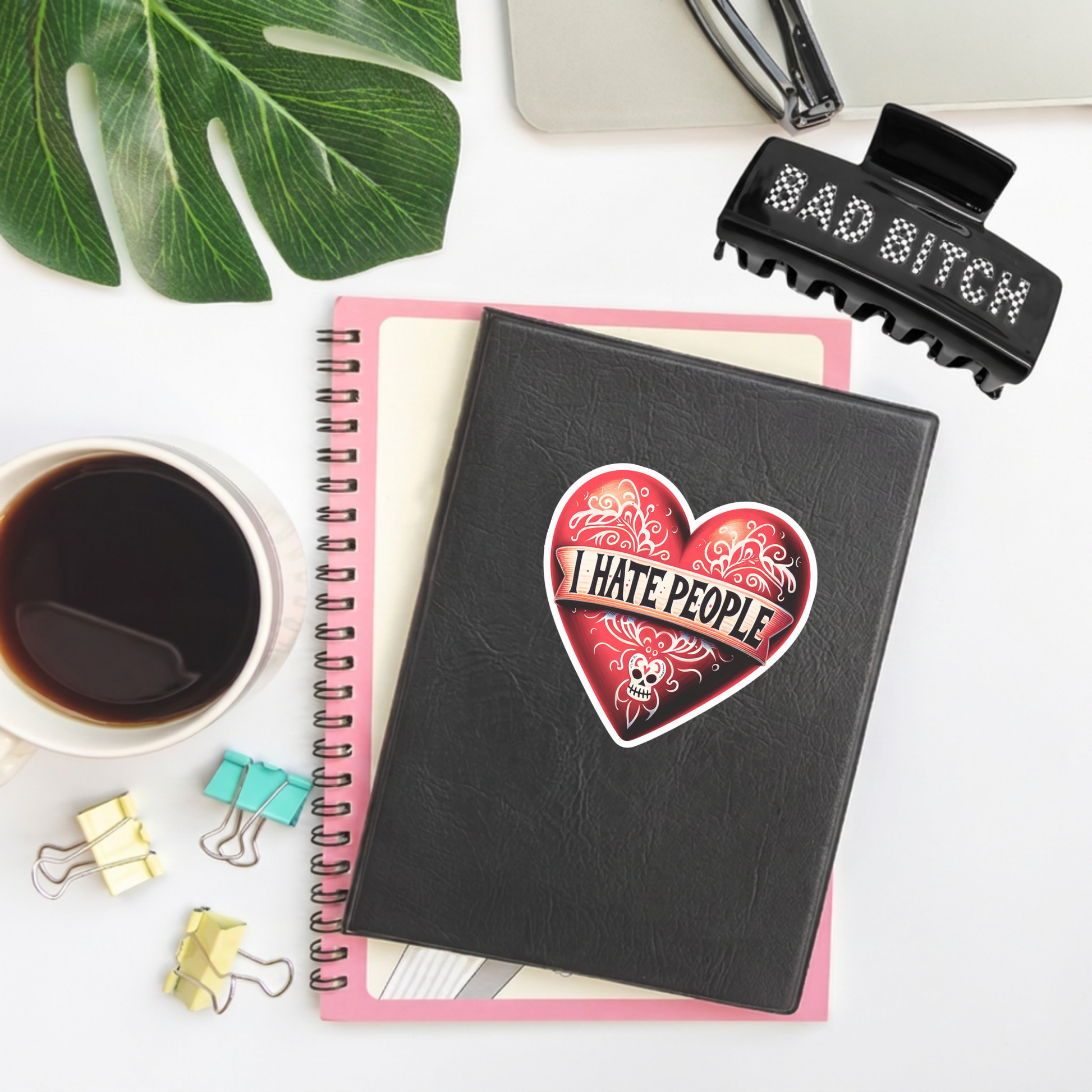 A black spiral notebook with a red heart-shaped sticker that reads 'I HATE PEOPLE' in the center, styled in a flat-lay with a coffee cup, binder clips, a green leaf, and a black hair claw clip on a white background.