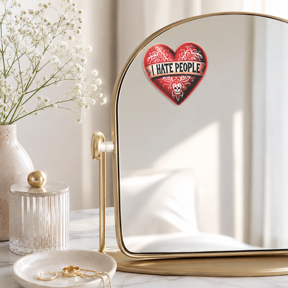 A gold-framed vanity mirror on a marble surface, reflecting a red heart-shaped sticker with the text 'I HATE PEOPLE' and a skull graphic. The scene is styled with a vase of white flowers, a glass jar, and a dish with gold jewelry in a bright, neutral room.
