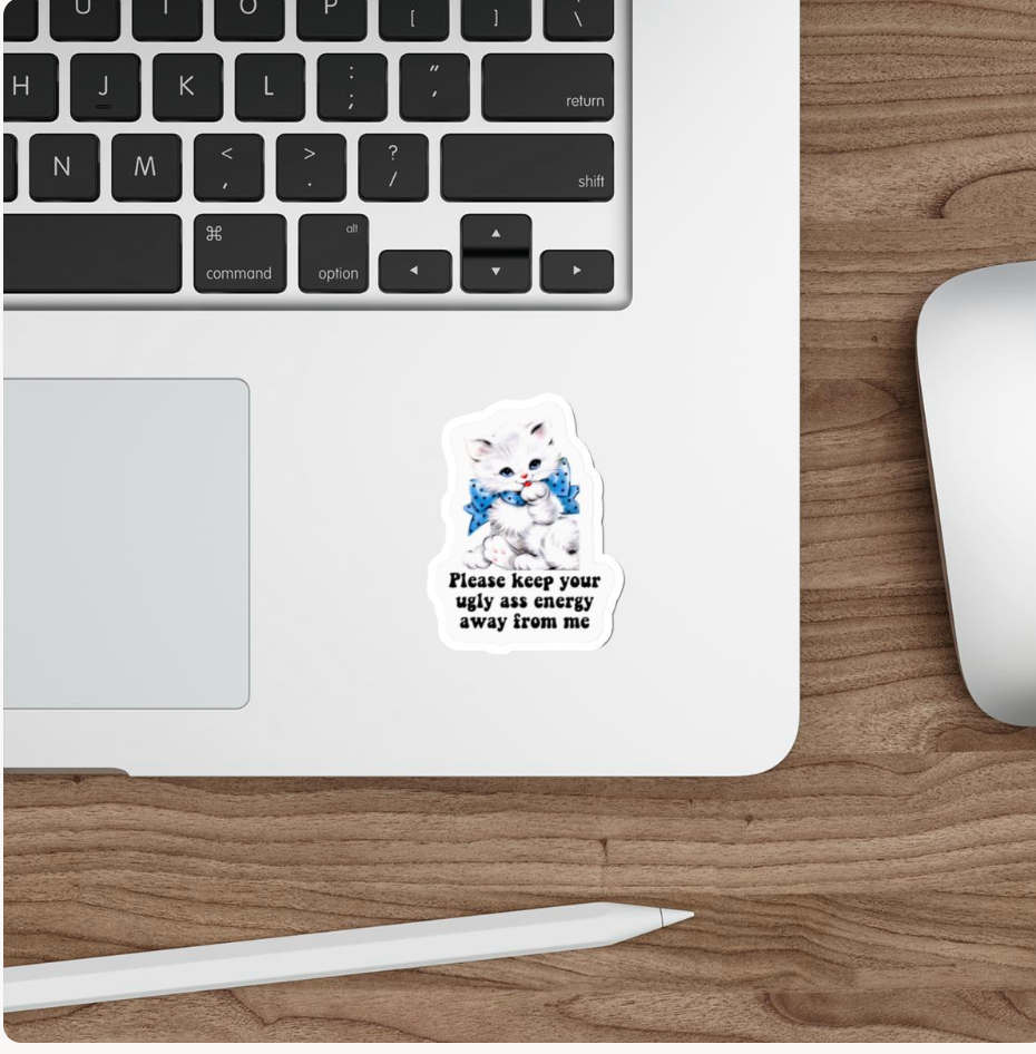 A die-cut sticker featuring a white kitten wearing a blue bow tie with the text 'Please keep your ugly ass energy away from me' applied to the bottom edge of a silver laptop. The laptop is shown on a wooden desk alongside a white stylus and a white computer mouse.
