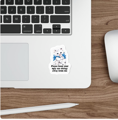 A die-cut sticker featuring a white kitten wearing a blue bow tie with the text 'Please keep your ugly ass energy away from me' applied to the bottom edge of a silver laptop. The laptop is shown on a wooden desk alongside a white stylus and a white computer mouse.