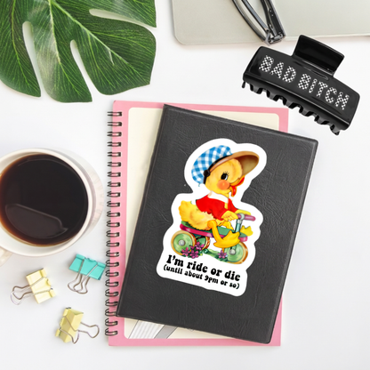 A black spiral notebook with a colorful cartoon duck riding a yellow tricycle sticker on the cover, styled in a flat-lay with a pink page insert, a coffee cup, binder clips, a green leaf, and a black hair clip on a white background.