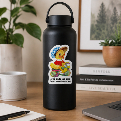 A matte black insulated water bottle with a carry loop stands on a wooden desk in a styled indoor setting. The bottle has a colorful cartoon duck sticker on the front wearing a blue-and-white checkered hat and riding a yellow bicycle, with the text 'I'm ride or die (until about 9pm or so)'.