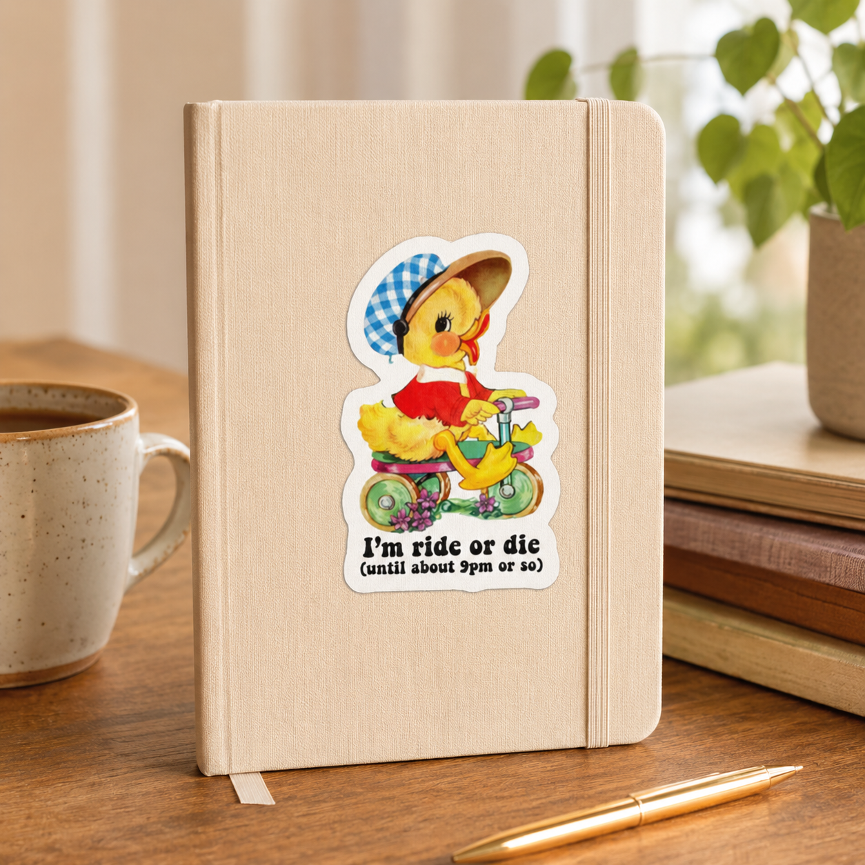 A beige hardcover notebook with an elastic closure band stands on a wooden desk. The front cover features a die-cut sticker of a cartoon duck riding a yellow tricycle, with the text 'I'm ride or die (until about 5pm or so)'.