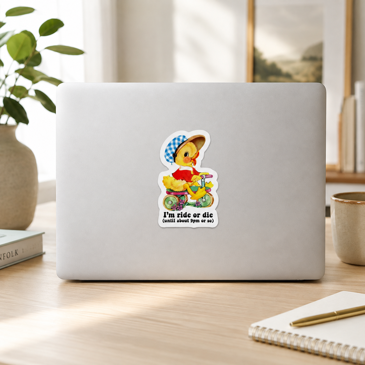 A die-cut sticker featuring a cartoon duck riding a yellow scooter with the text 'I'm ride or die (GOD SAVE TYT ANO M)' shown on the back of a silver laptop, placed on a wooden desk in a bright, modern room.