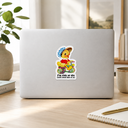 A die-cut sticker featuring a cartoon duck riding a yellow scooter with the text 'I'm ride or die (GOD SAVE TYT ANO M)' shown on the back of a silver laptop, placed on a wooden desk in a bright, modern room.
