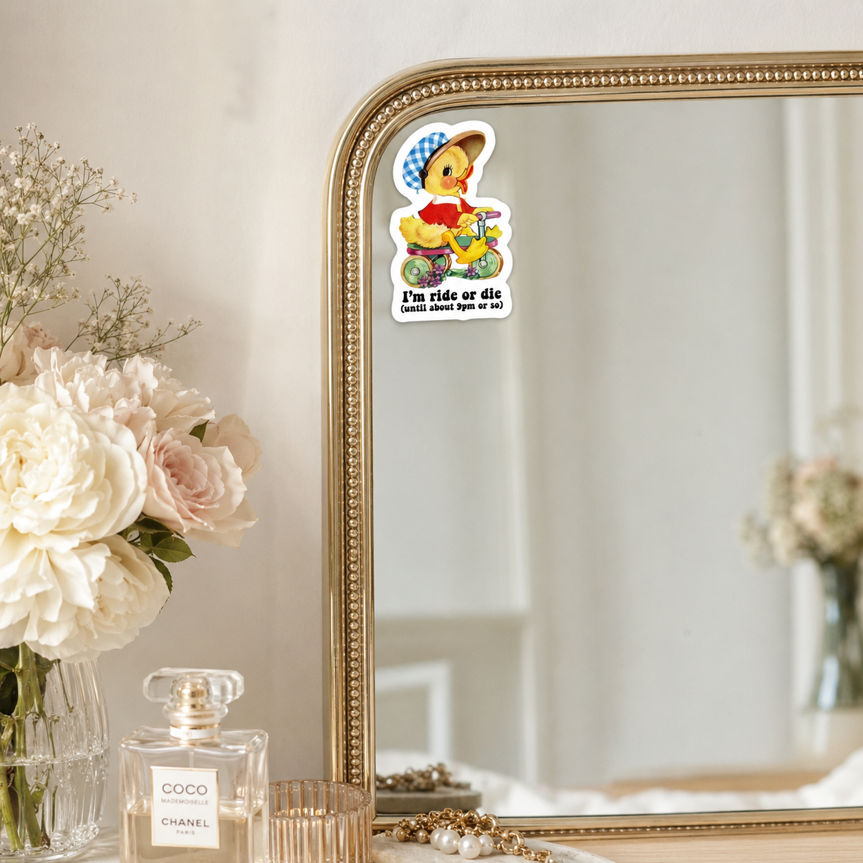 A die-cut sticker featuring a cartoon duck on a bicycle with the text 'I'm ride or die' is applied to the top edge of a gold-framed mirror. The scene is styled with a vase of white and pink flowers and a Chanel perfume bottle on a tabletop.