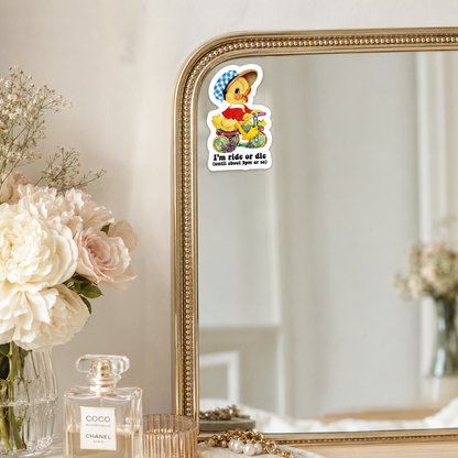 A die-cut sticker featuring a cartoon duck on a bicycle with the text 'I'm ride or die' is applied to the top edge of a gold-framed mirror. The scene is styled with a vase of white and pink flowers and a Chanel perfume bottle on a tabletop.